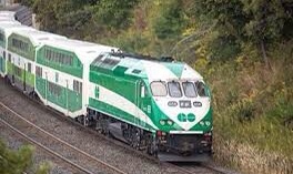 A green & white GO train zooms through a forest, heading somewhere full of people.